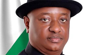 Public figure in formal attire with a black shirt and hat, standing in front of a blurred Nigerian flag background.