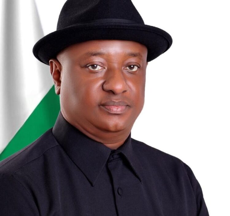 Public figure in formal attire with a black shirt and hat, standing in front of a blurred Nigerian flag background.