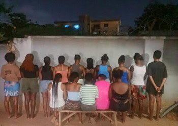 A group of people gathered outdoors at night, sitting and standing along a white wall, in a community setting.