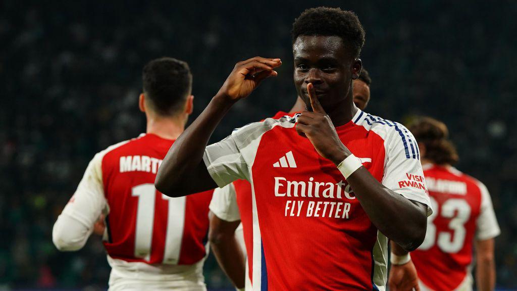 A vibrant Arsenal football player celebrates on the pitch, holding a finger to his lips in a 'shh' gesture, with teammates in the background during a match.