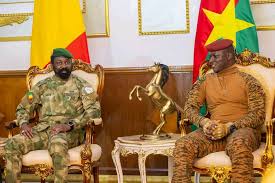 Soldiers in formal attire seated on ornate chairs in a formal setting with flags in the background.