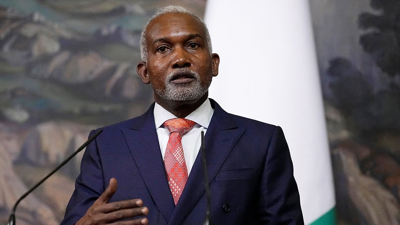 Senior man in formal suit speaking at a press conference with a microphone and a national flag in the background, promoting leadership and governance.