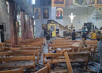 Ruined church interior with damaged wooden pews, fallen debris, and worship paintings after an attack, with visitors inspecting the destruction during daylight.