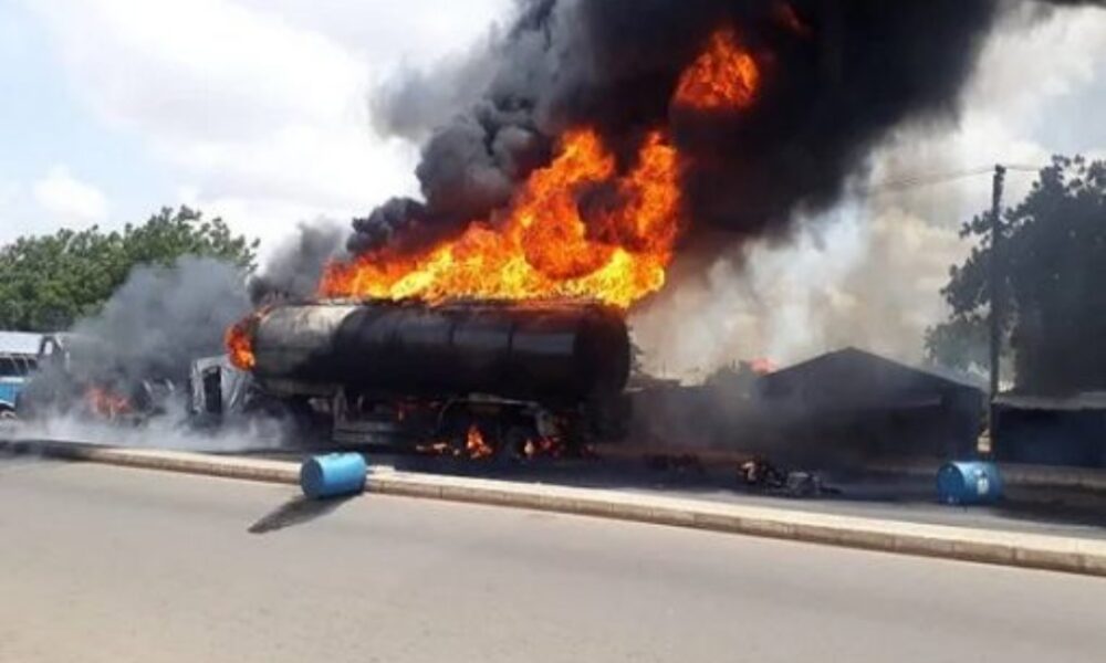 Exploding fuel tanker on the road, flames and thick black smoke, emergency situation, urgent response needed, danger of fire spreading, roadside accident, disaster scene, safety hazards, vehicle combustion, freight transport incident, crisis management.