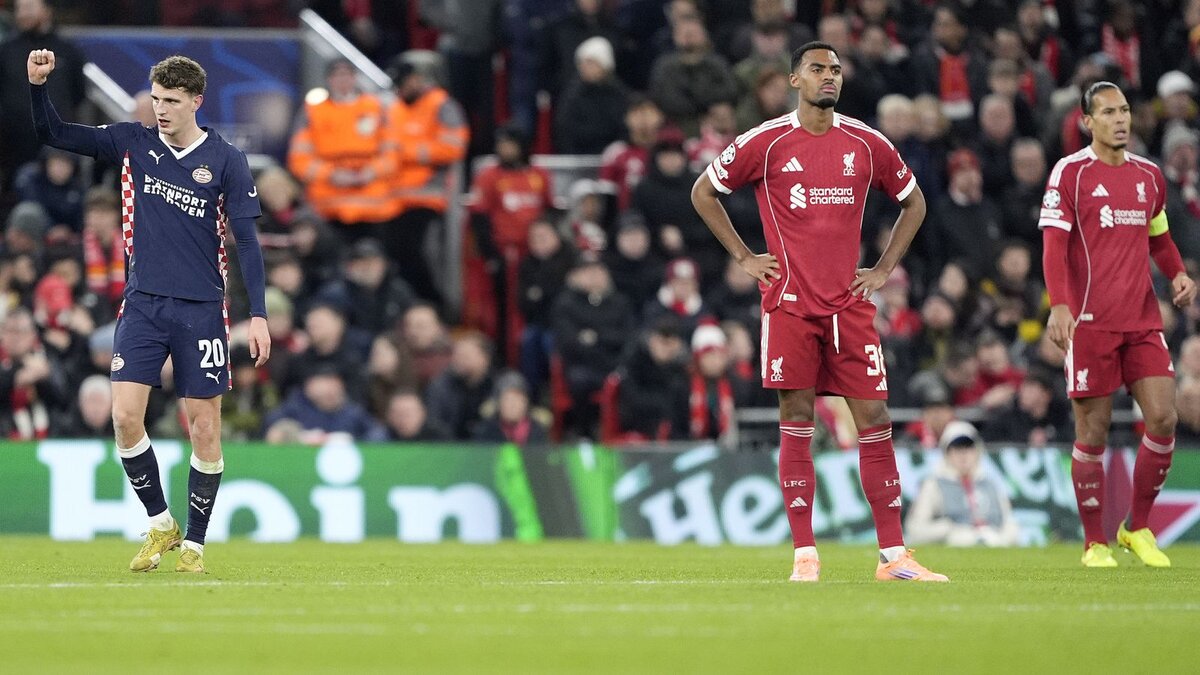 A football match scene showing players in Liverpool and PSV Eindhoven kits on the pitch with a crowded stadium background.